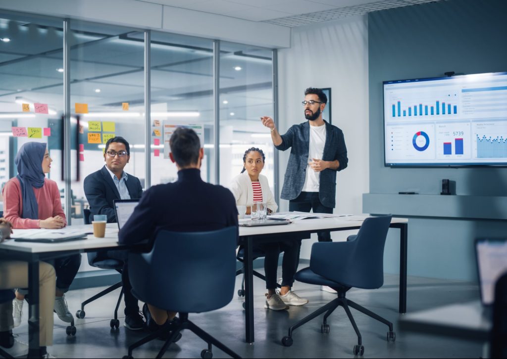 Diverse Modern Office: Businessman Leads Business Meeting with Managers, Talks, uses Presentation TV with Statistics, Infographics. Digital Entrepreneurs Work on e-Commerce Project.