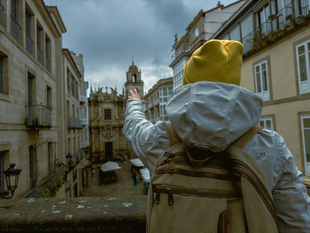 solo traveler clad in a white jacket and a yellow beanie waves towards the historic architecture